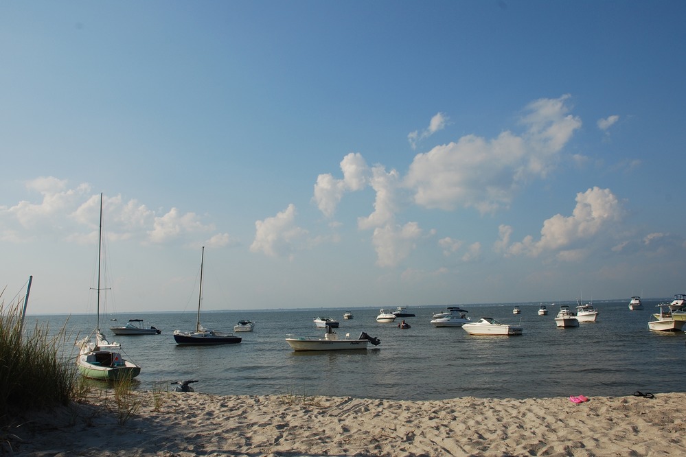 At Fire Island National Seashore's Talisman/Barrett Beach site, small boats lie quietly anchored in a calm bay, under a clear blue sky embellished with puffy white clouds.
