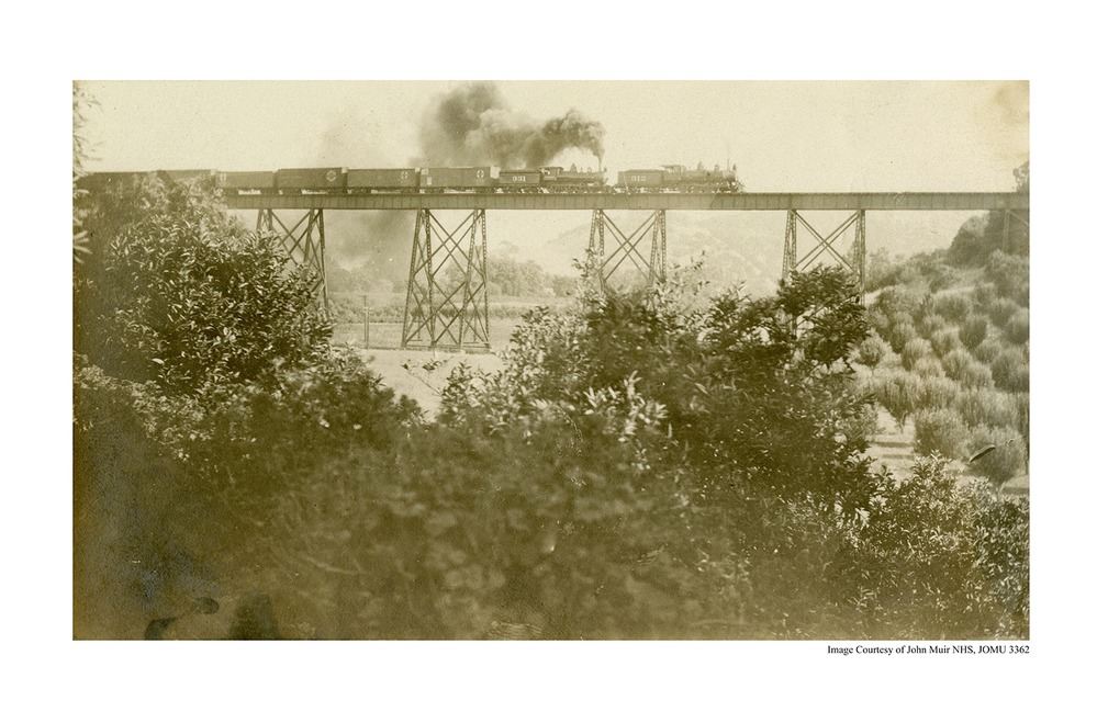 A historic photo of a railroad trestle can be seen in the background. A train with stack smoke can be seen moving along it. Trees are in the foreground. 
