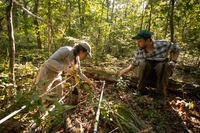 Two biologists kneel next to a quad, which is white PVC pipe in the shape of a rectangle and look at the seedlings within it's boundaries