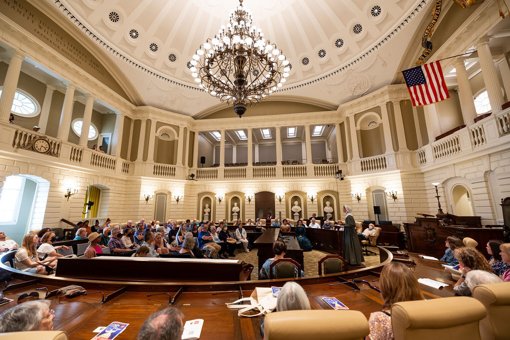 Circular room with a gallery. On the main floor are two semi-circles of desks with people sitting in them. Above is a chandelier.