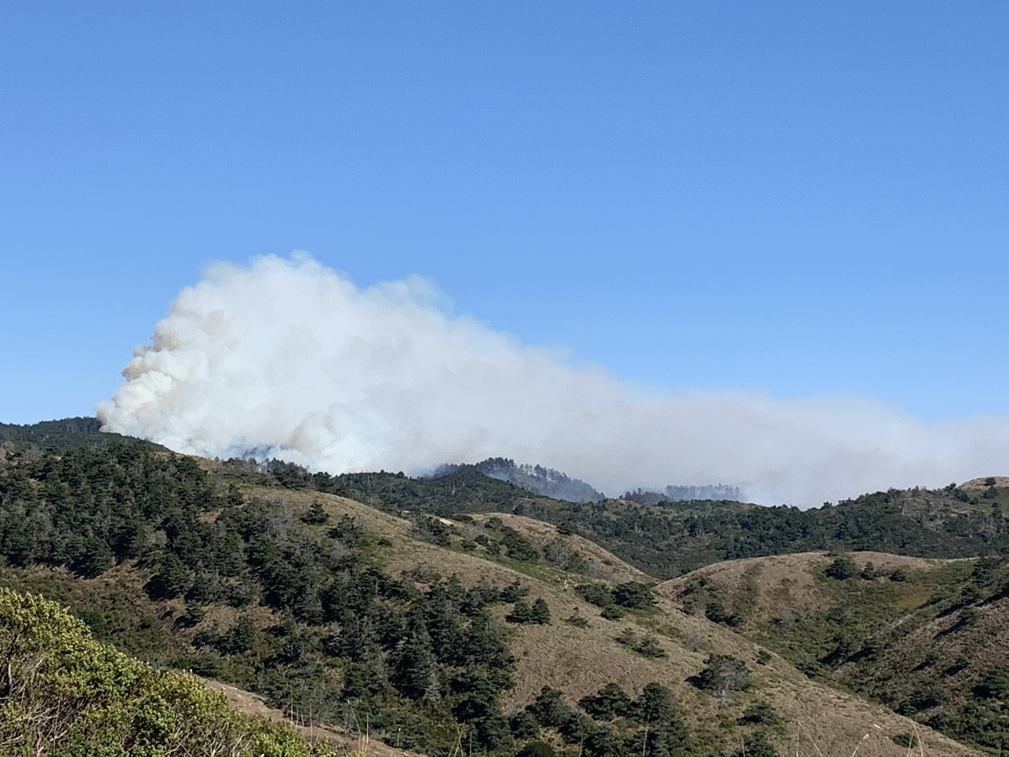 A thick plume of smoke rises into cloudless skies from a forested ridge in the distance before being blown to the right along the ridge crest. Sparsely wooded ridges are in the foreground.