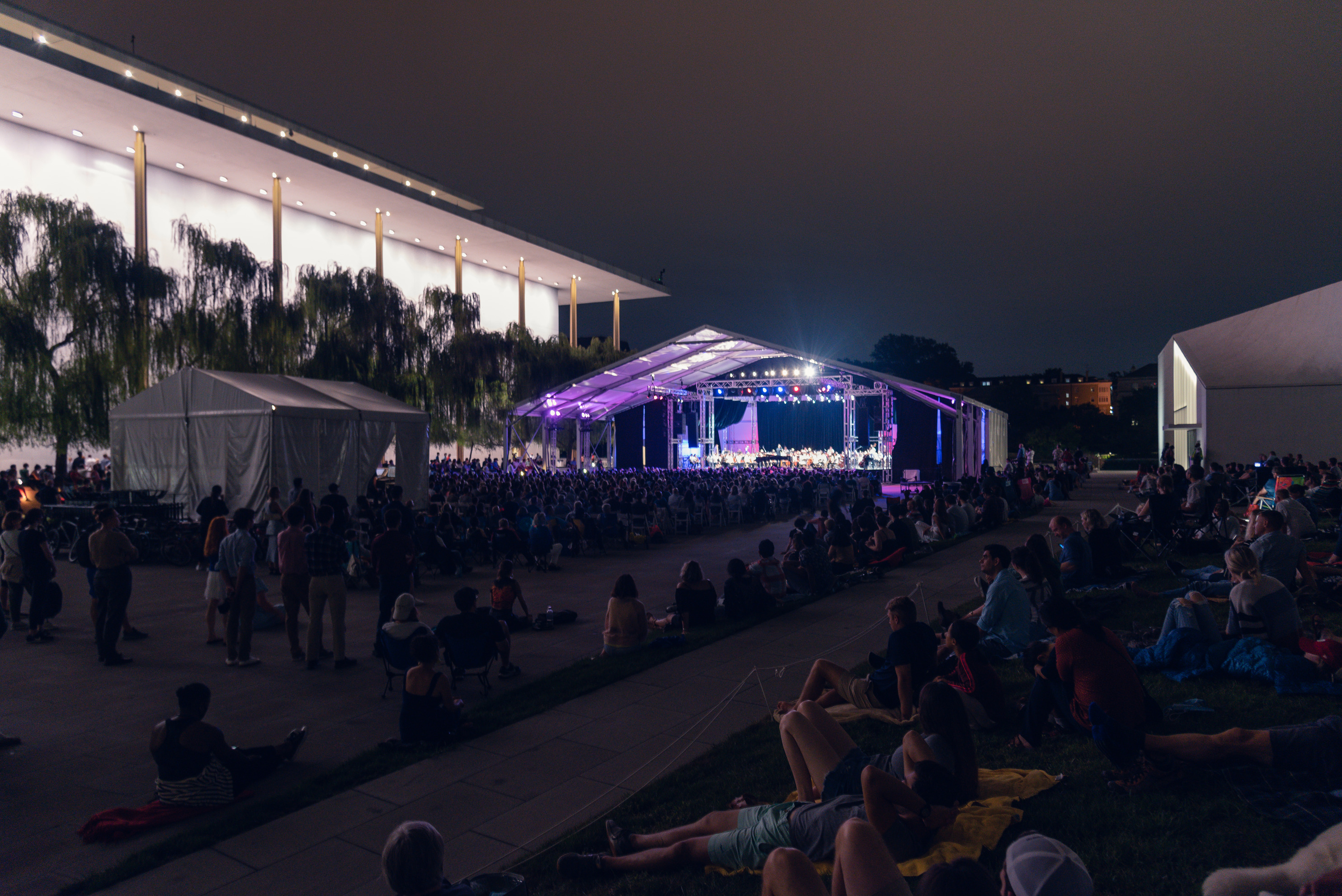 A crowd is seated under a large balcony far from the stage and performance of the National Symphony Orchestra.