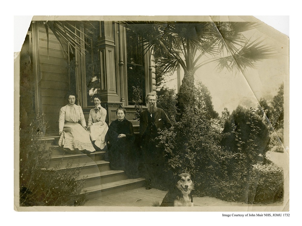 An historic photo of two younger women in dresses, an older woman in a dress an older man in formal dress and a dog can be seen sitting on the porch steps of the Muir home. Trees and plants are seen the photo.