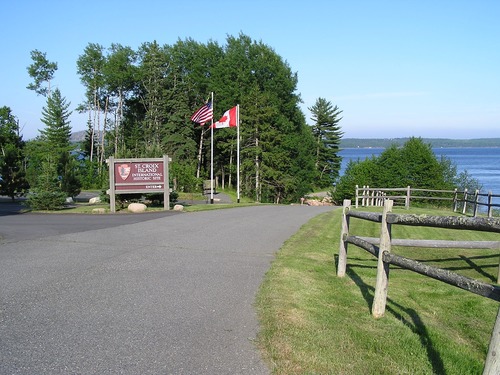 A road leads to an ocean view with tall trees. On the right of the road is a grassy area with a wood fence. On the left are flag poles, one with the United States flag and one with the Canadian flag. The flag poles are next to a large brown, illegible sign with the National Parks arrowhead logo.