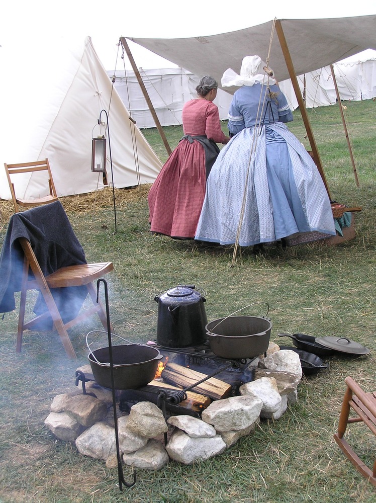 Two women with their backs turned toward the camera as they work on some food preparation while two Dutch ovens and a coffee pot heat up over a campfire.