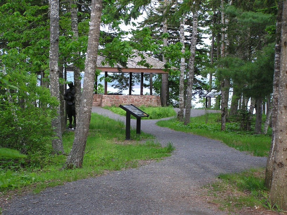 An asphalt path leads through trees, past two wayside exhibits and a bronze statue toward a pavilion.