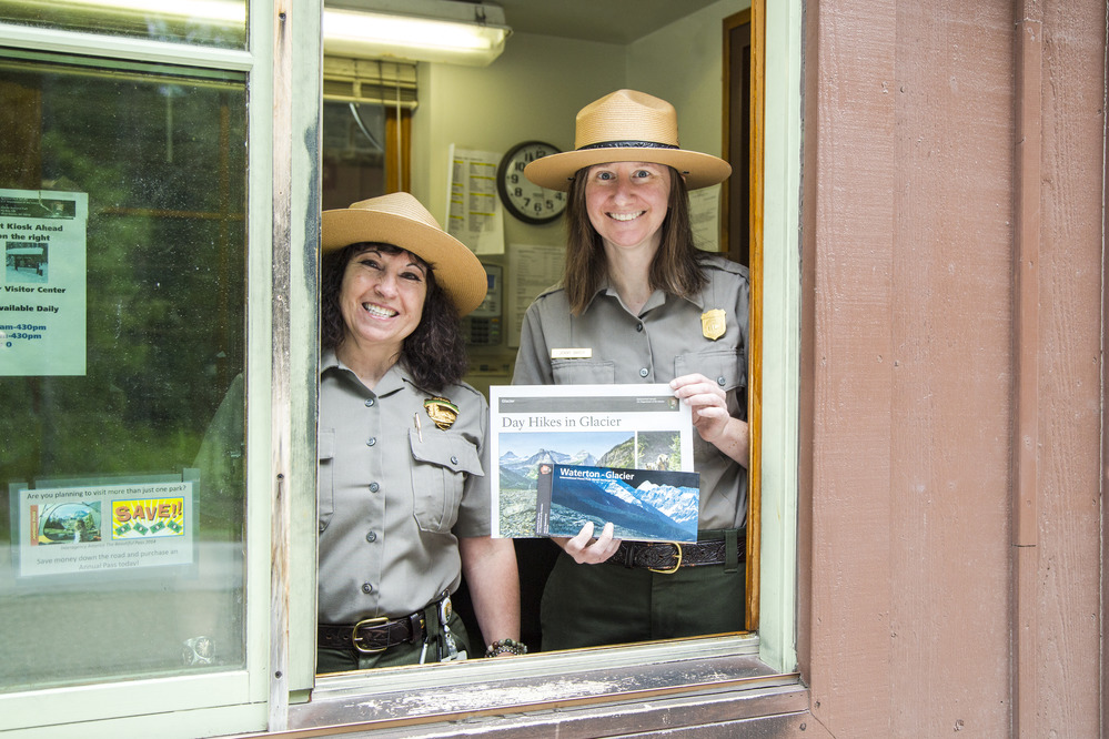 Two rangers in a fee booth window with one holding a park brochure and newspaper