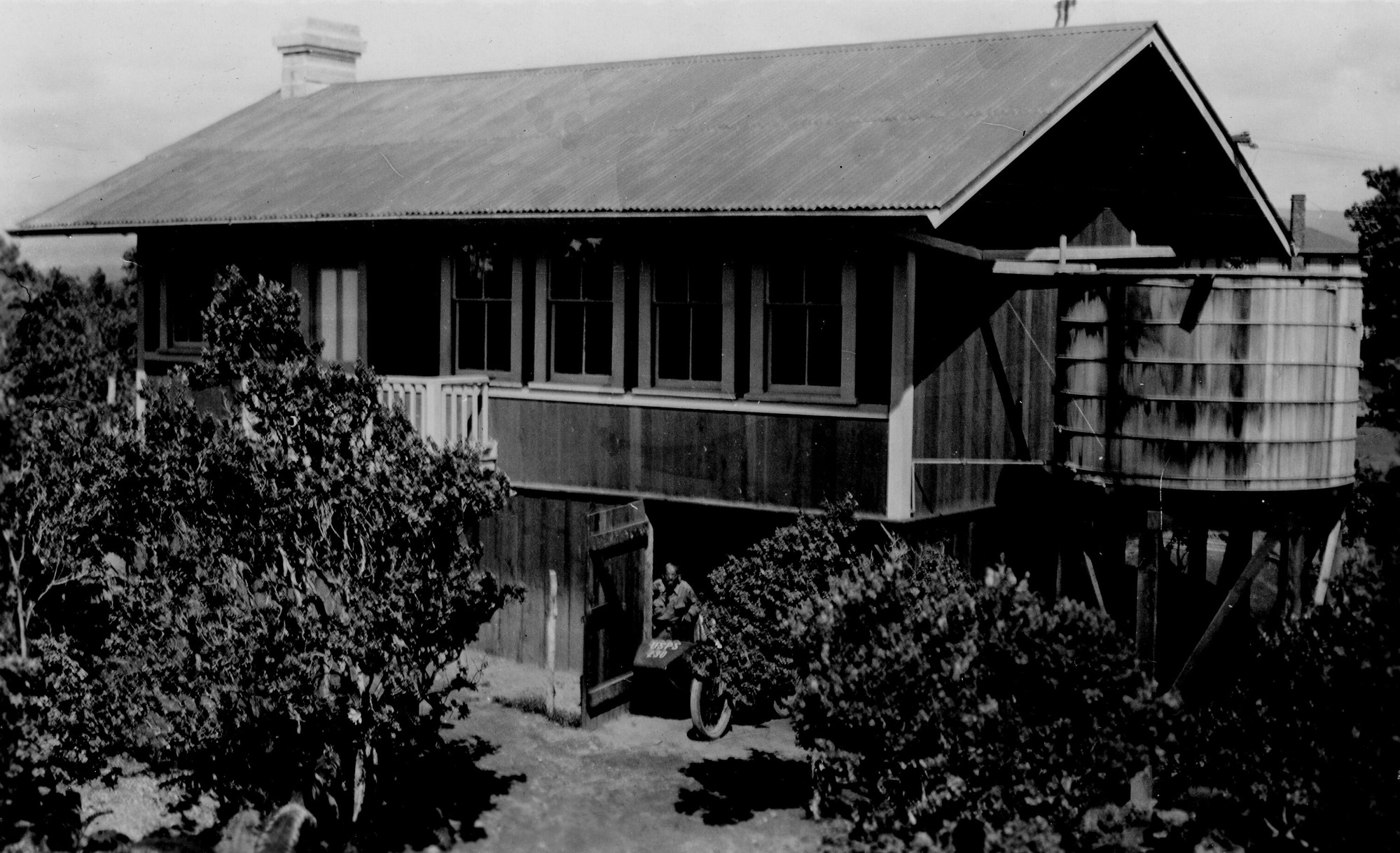 Black and white photograph of the back side of a two-story building. A catchment tank sits directly next to the building’s right side, closest to the camera. Six windows are aligned horizontally along the back wall. Lush vegetation is present around the house. A man is visible on the ground floor through an open door. It appears he is next to a bike which reads “USPS 230” on its front.