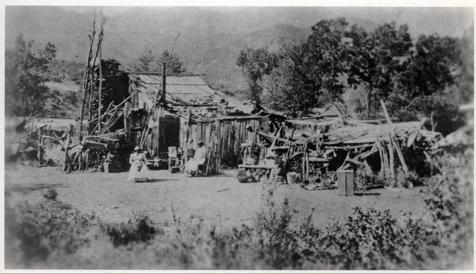 Two women sit outside a small wooden house with rock chimney and sloping roof with a mountain range in the background.