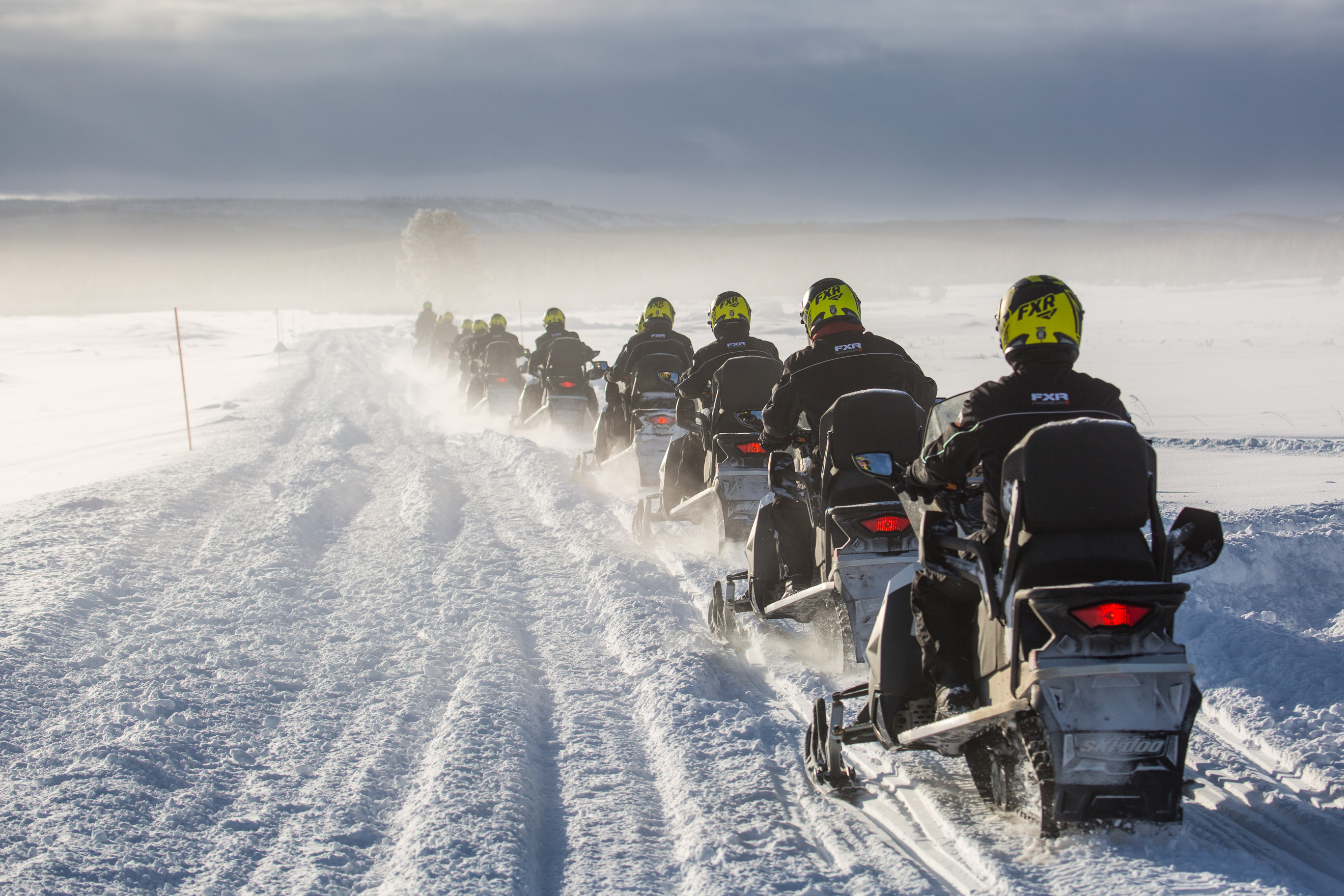 Line of snowmobilers driving away from camera on a road on a snow covered flat