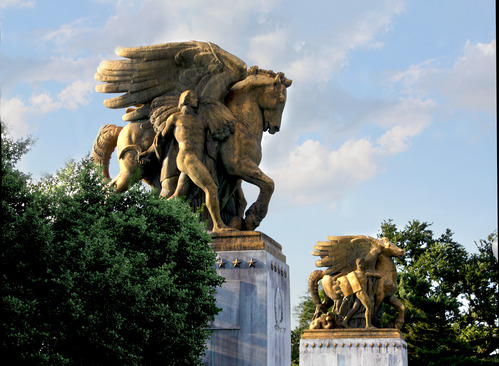 Two golden sculptures of winged horses with men standing beside them. 