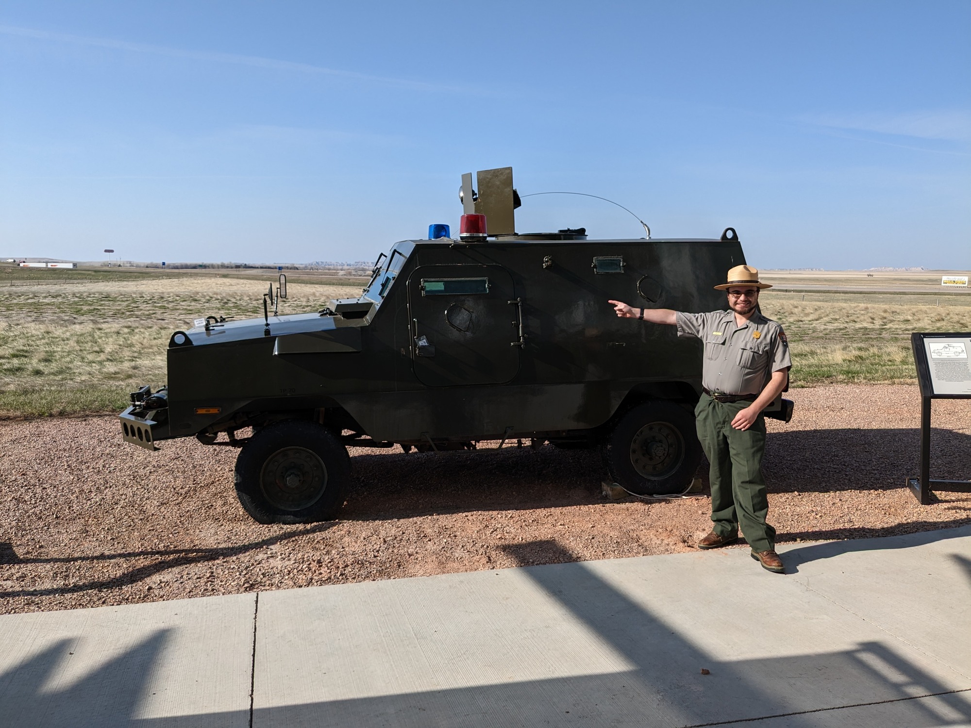 Man in park uniform of gray shirt, green pants, and straw flat hat pointing at a green armored truck outside on a gravel pad with prairie behind it. Brown hair, glasses, and mustache, and glasses. 
