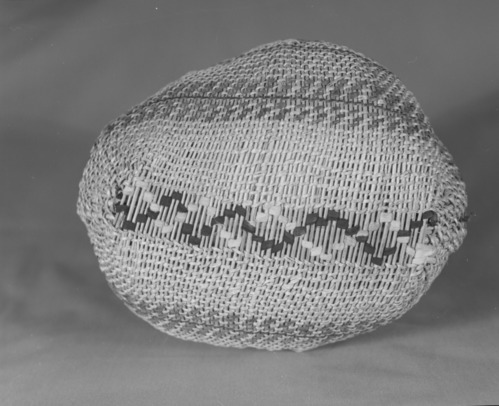 Private Collection. Object: Miwok basket. Dimensions:H-8" x L-9" x W-7 1/2" (maximum) mouth 5". Materials: Scraped shoots, split shoot (probably Ceanothus cuneatus), redbud, bracken fern root, black cloth & red cloth (now white). Technology: Cradle board hood start utilizing cloth wefts. Body of basket in spaced rows of diagonal twining 1/8 to 1/4" apart, one row three strand twining one inch below selvedge, 3 short rows of twining apparently used as a filler on one end of basket. Rim finish of rods folded to left and coiled with small bundle of rods coiled in a 2" section to even out contour. Rim stick attached with aid of an awl. Collection History: From collection of Florence Hill McCullough, daughter of Thomas Hill purportedly from Hill's studio. Photographed by Michael Dixon. Catalogued by Craig Bates and Barbara Beroza. Yosemite Collections, National Park Service.