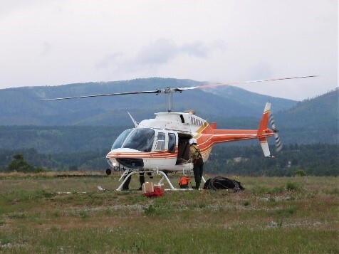 A white and red helicopter is landed in a grassy, green field with the pilot loading supplies into the open door of the main compartment