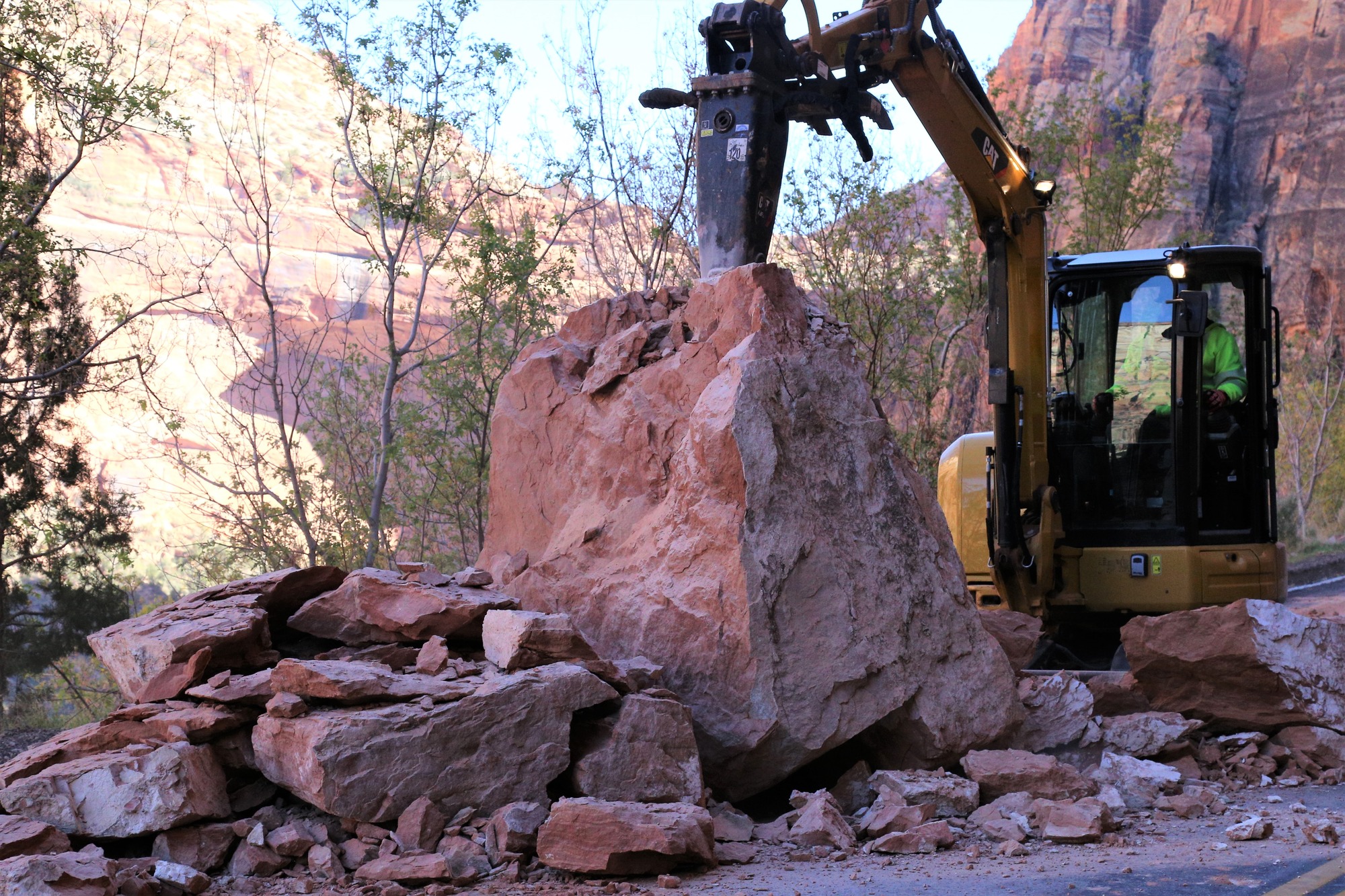 A large boulder with smaller pieces falling off from it, behind it an excavator with a jackhammer attatched picking away at the rock.