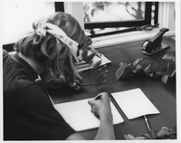 A black and white image of a man drawing or taking notes of a plant specimen. The man is located on the left side of the image. He is looking down at the paper in front of him. He is wearing a bandana and a stripped polo shirt. He is holding a pen in his right hand and is writing on a piece of paper. Also, he is holding a plant specimen in his left hand. The plant has short branches with bristle-like leaves. On the desk there is a stapler, a clipboard, and other plant specimens. In the background a double pane window is partially open.