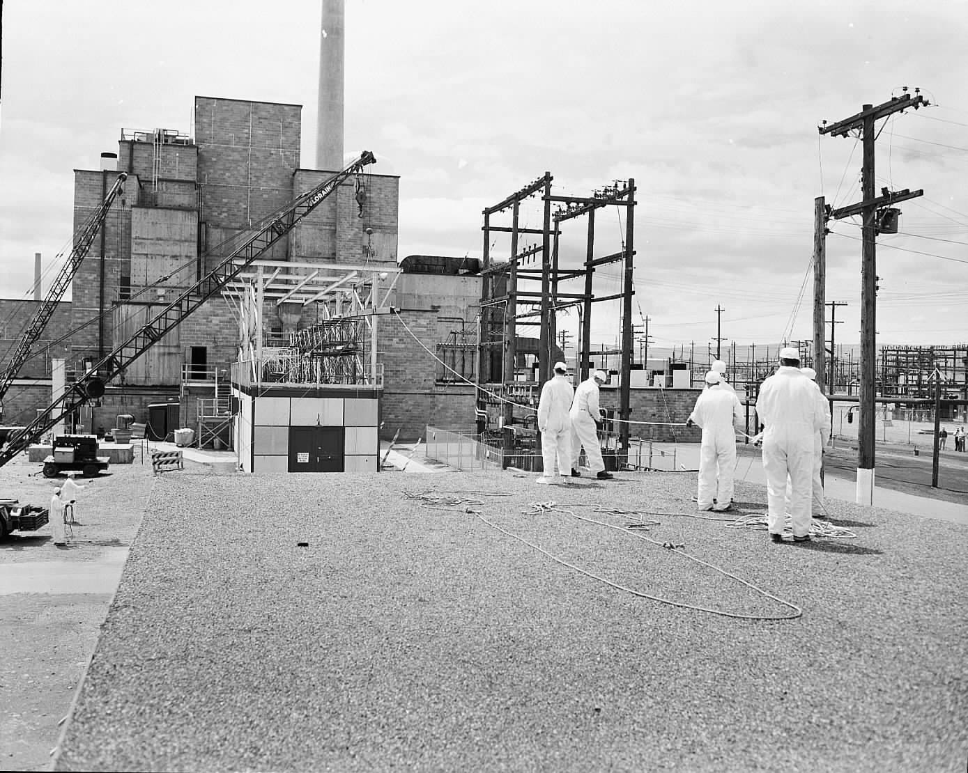 Black and white photo of several people in white uniforms and caps in front of a blocky, large building with a ventilation stack. 