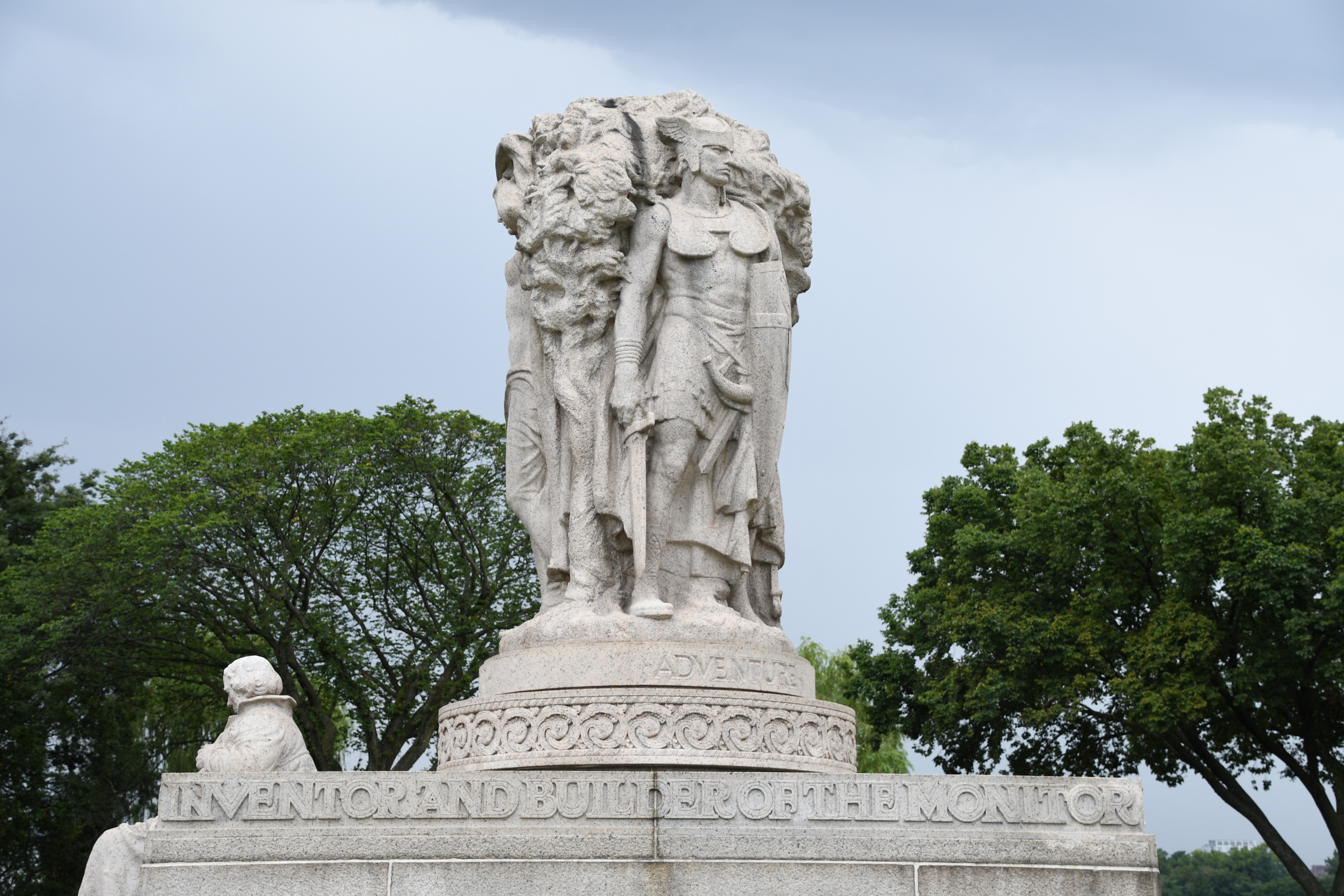 Another side of the granite memorial depicting a man with the words "adventure" below, the circular pattern design surrounds the statue, and below it reads "inventor and builder of the monitor." 