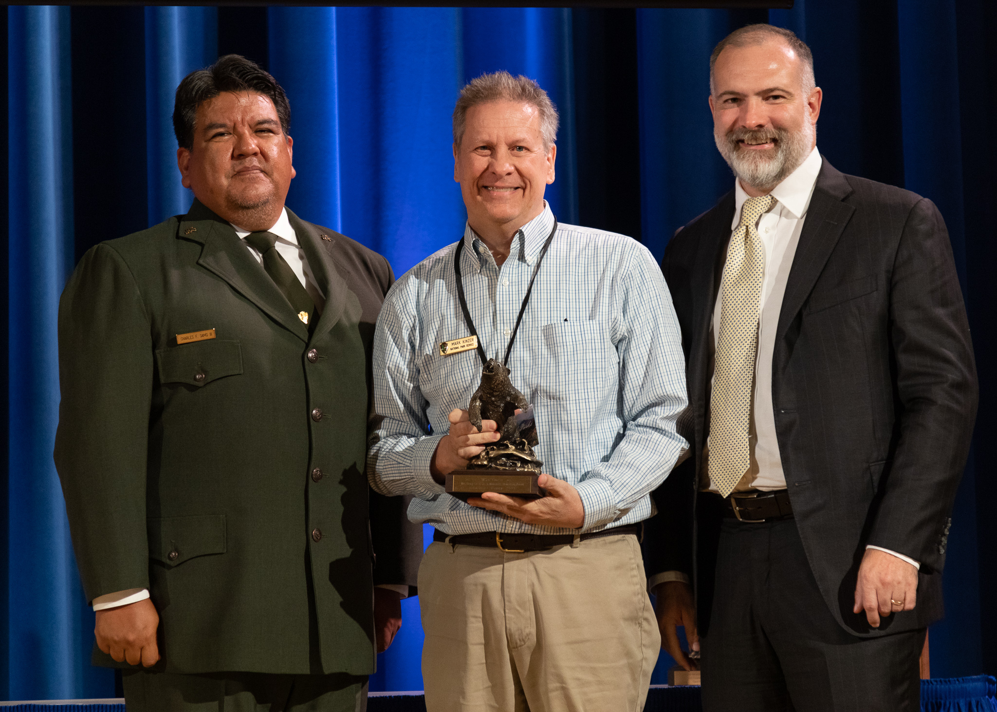 A man accepts an award in between the Director of the National Park Service and Deputy Secretary of the Department of the Interior.