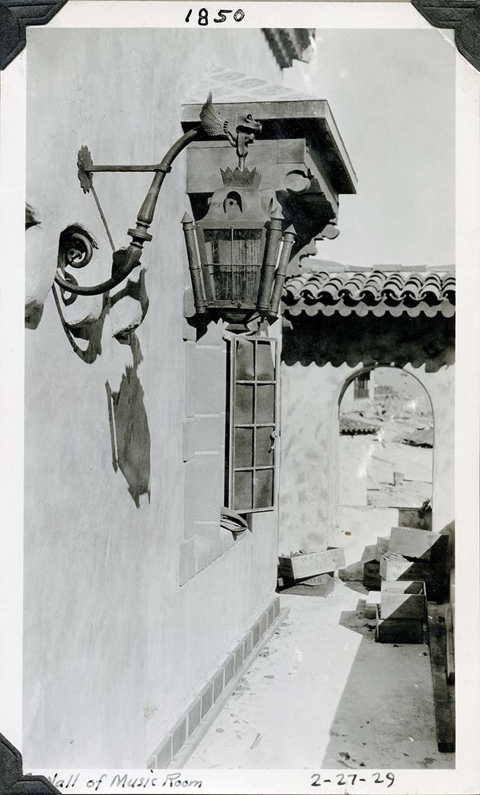 This is an historic black and white photograph from the Scotty's Castle Historic Photograph Collection, Death Valley National Park of elaborate hand wrought metal lantern mounted on hand wrought bracket on exterior stucco wall. Lantern base four sided with fitted top. S-shaped bracket has dragon like hook for lantern. Stack of wooden crates stamped: W P Fuller. Background shows narrow eight pane window open. Ends with arched wall supporting small red-tiled roof. Inscriptions in black ink along upper and lower border.