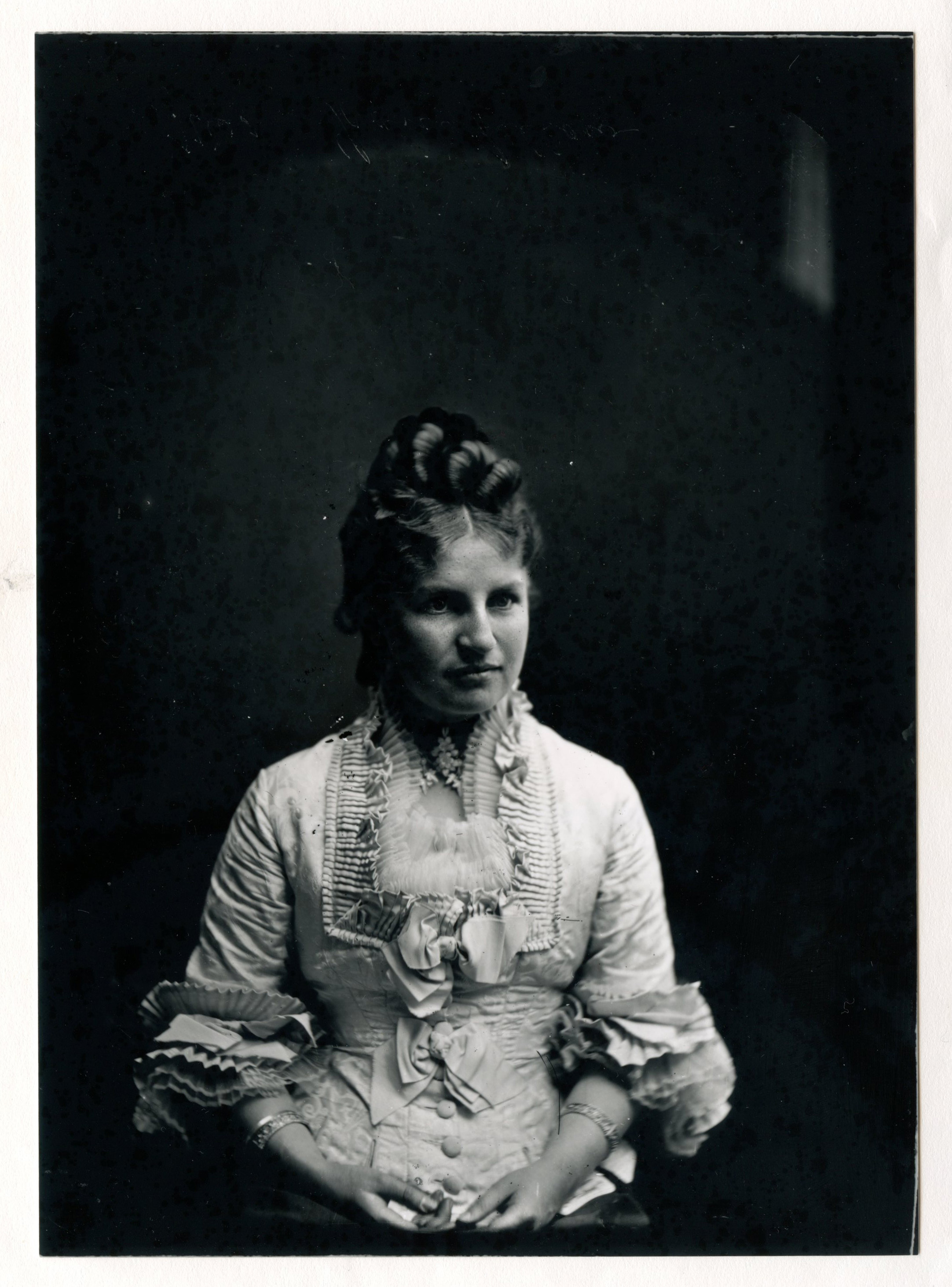 A young dark-haired white woman sits with her hands in her lap. She is wearing a dress with many ruffles.