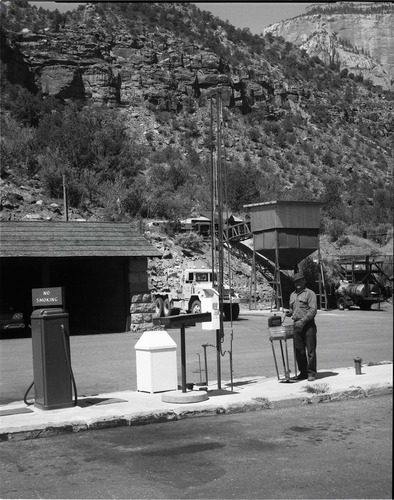 BW Photos of man standing next to maintenance equipment. Vehicle fuel pumps in foreground, equipment and dry sand hopper in background.