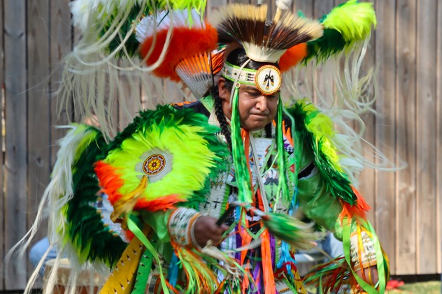 An Alabama-Coushatta dancer wearing brightly-colored regalia, featuring bright green, dark green, orange, red, and white feathers.