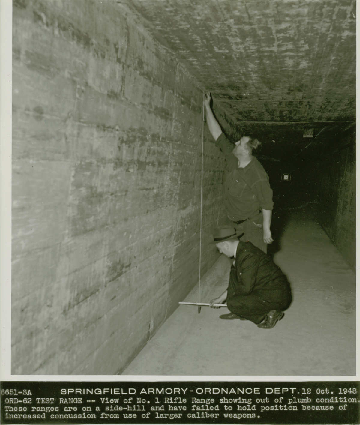 Black and white photo of two men in a narrow hallway. One man holds the top of a string to the ceiling. The other man is on the ground with the stick that is attached to the end of the string. Text under the photo reads, “6651-SA Springfield Armory – Ordnance Dept. 12 Oct. 1948, ORD-62 TEST RANGE -- View of No 1 Rifle Range showing out of plumb condition. These ranges are on a side-hill and have failed to hold position because of increased concussion from use of larger caliber weapons.”