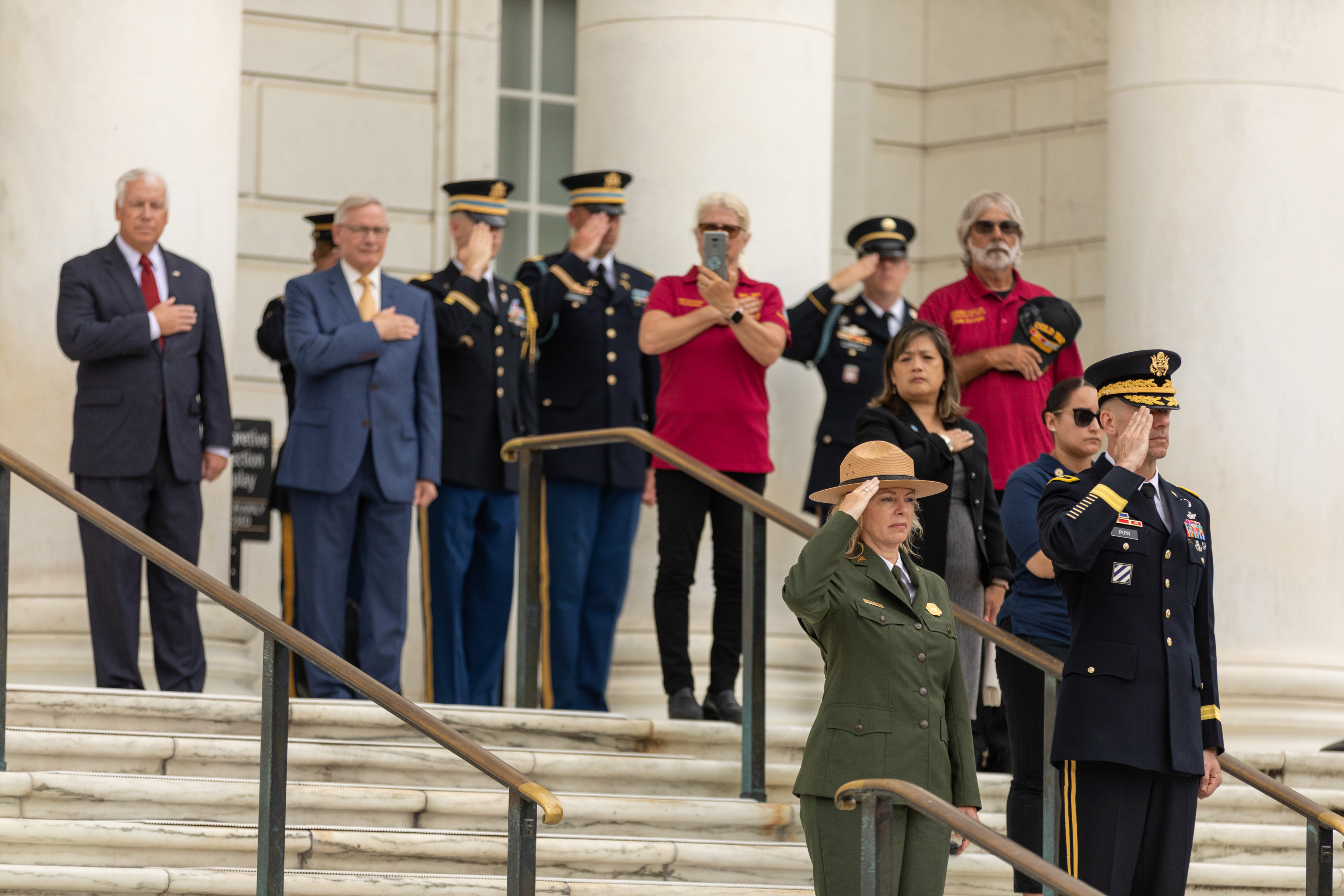 Director Hall and General Pepin stand on white marble stairs and salute. Behind them other military personnel and NPS staff salute or hold their hand over their hearts.