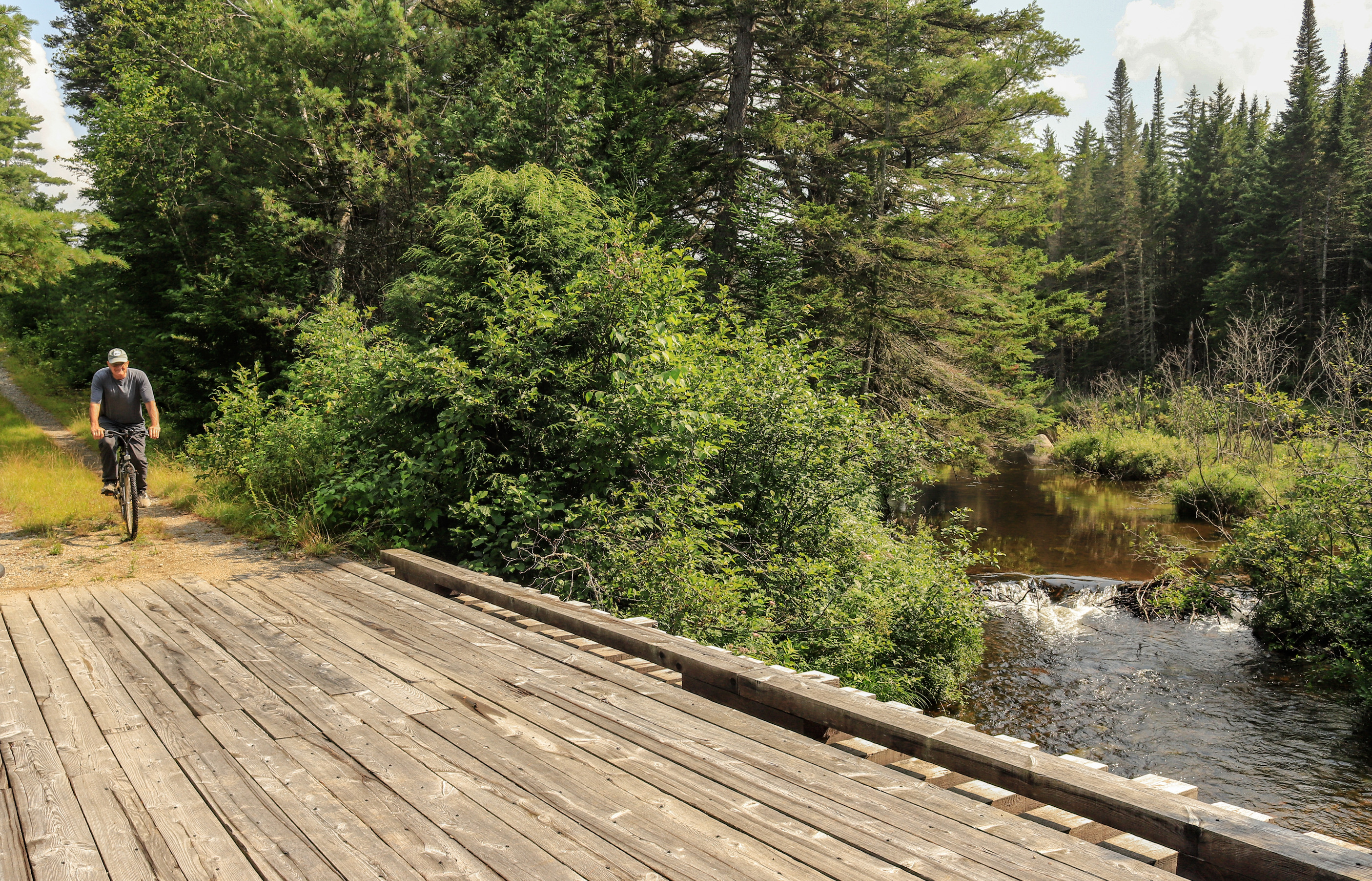 A wide angle image with a cyclist coming down the path on the left side and a bridge and stream in the foreground of the right side