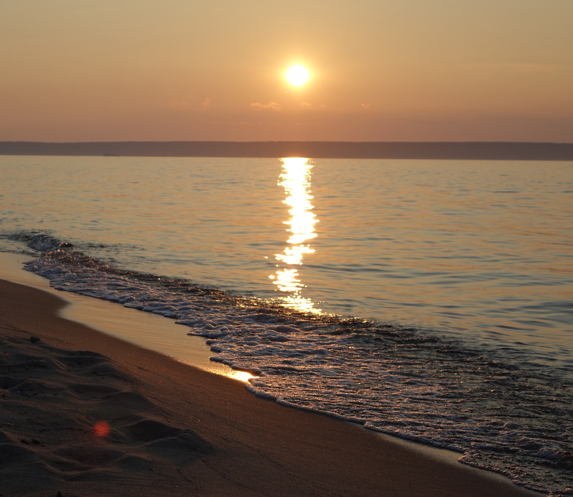 Sun is nearing the horizon over Lake Superior. The sun reflects on the water in a long line towards the photographer.  Water laps onto the sandy shore in the foreground.