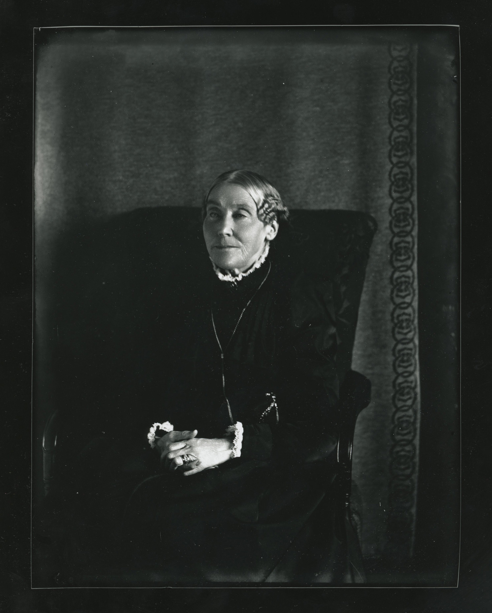Older white woman in dark clothing sits with her hands clasped in her lap in front of a drape backdrop.