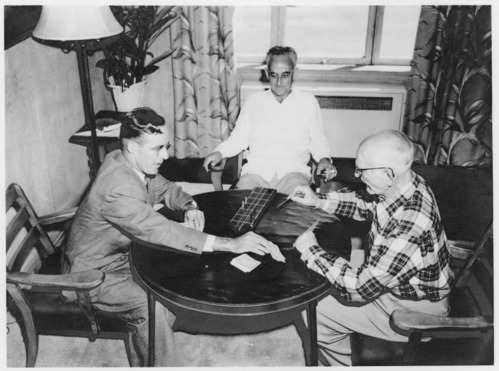 Black and white. Two men sitting at a table playing a card game. One man observing