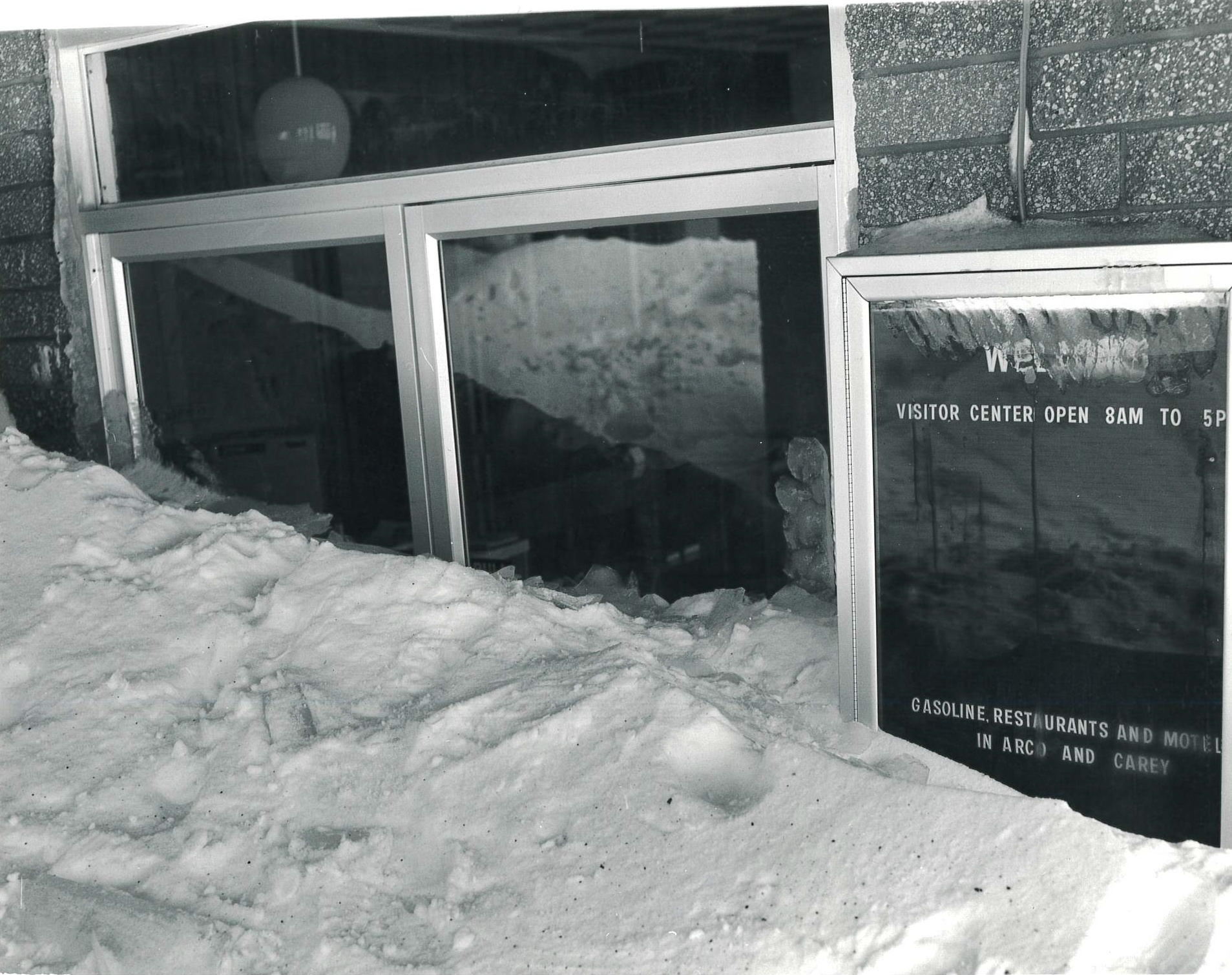 snowdrift covering more than half of a set of glass doors outside a brick building. a frosty glass bulletin board with visitor center hours is partially obscured by the snow.