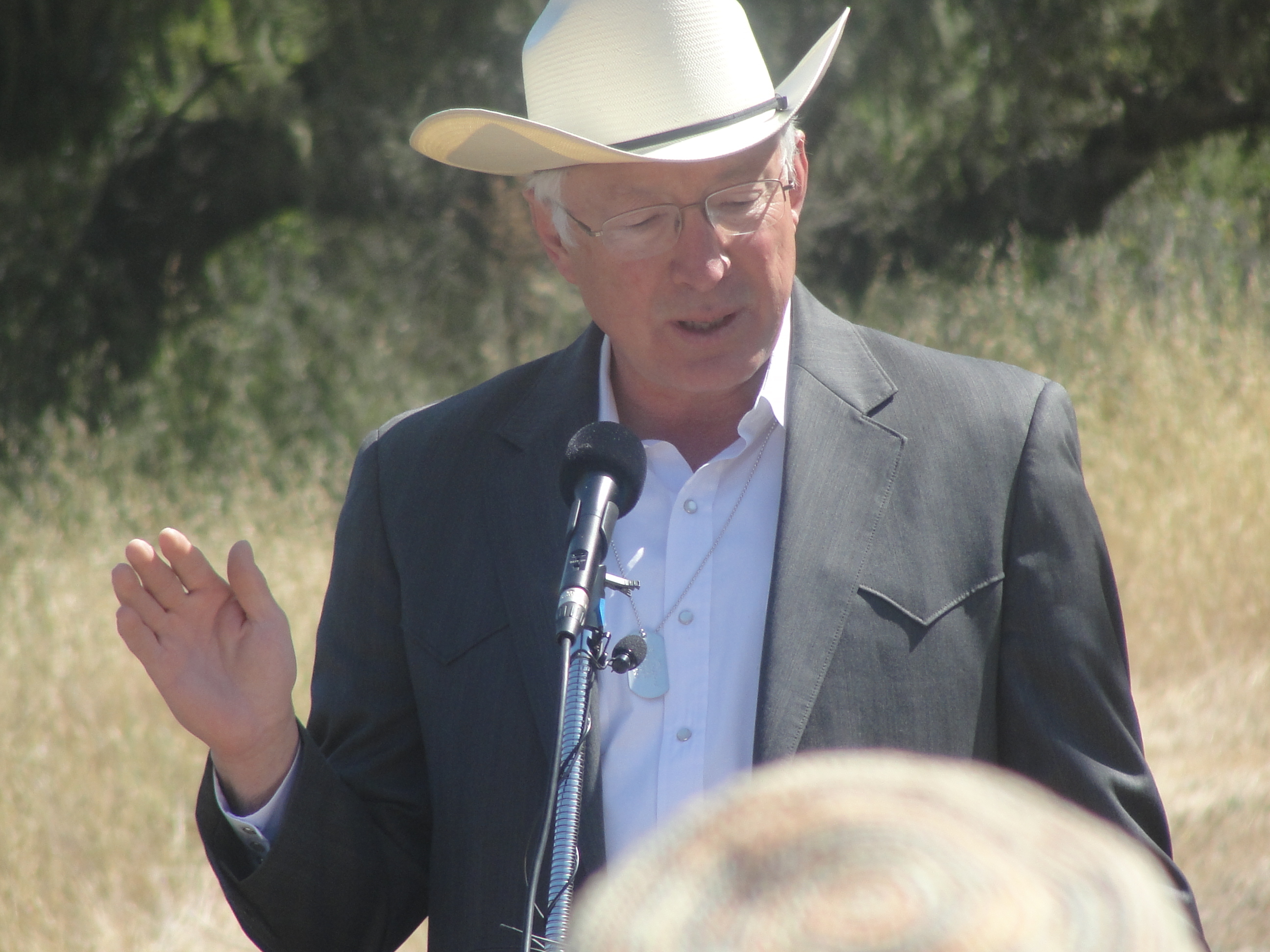 A person in a suit and cowboy hat stands at a podium outside and speaks