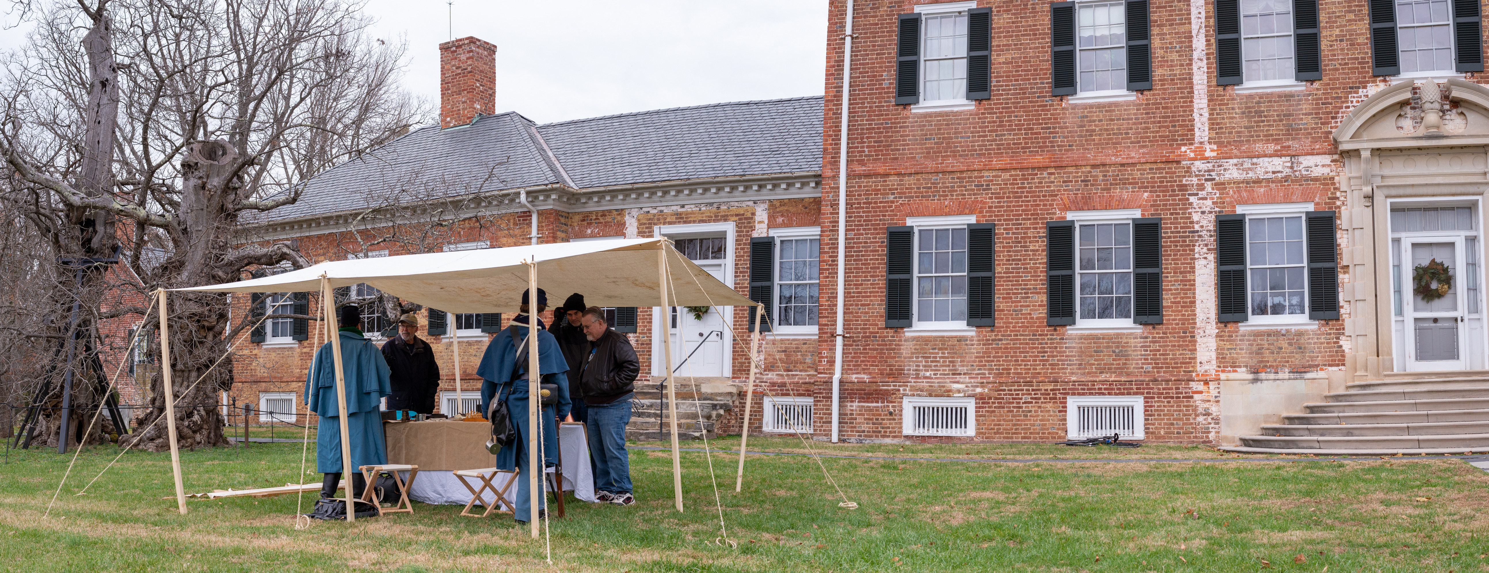 Two living historians dressed as a Union doctor and soldier stands at a table with medical supplies speaking to visitors with Chatham Manor in the background.