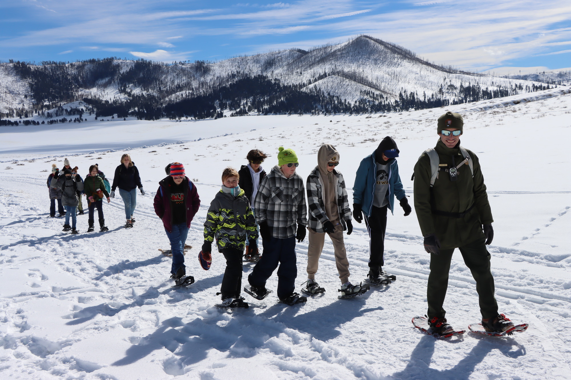 A park ranger leads a large group of students on a snowshoe hike in a mountainous landscape.