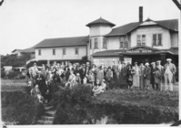 The image is in black and white. The focus of the picture is a large group of people posed in front of a hotel. In the foreground, there is an embankment with stairs leading up to the front grounds of the hotel. There are people sitting on top of the embankment and stairs, while the rest of the group stands along a path in the front gardens. Everyone is well-dressed, wearing coats and hats. The group spans from the right edge of the image to nearly the left edge of the photo. Directly behind the group is the hotel. A sign under the eaves of the front entrance's roof reads, 