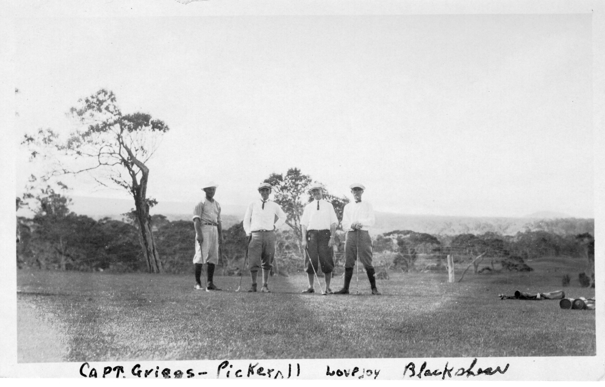 The image is in black and white. There are four men posed on the green of a golf course, left of the center of the photo. The men are standing in a row. All of the men look at the camera, wearing flat caps, shirts, trousers, socks, and shoes. The men are identified in the photo from left to right: Captain Griegs, Pickerall, Channing Lovejoy, and Blackshear.  The grass is short and sparse. Behind the men, a few large trees grow, including one to the left of the group. Past these trees and a wire fence, which stretches from one end of the photo to the other, is a heavily forested area. A hill is faintly visible far off in the distance on the right edge of the photo. Label at the bottom of the image in the white border of the photo reads, "Captain Griegs, Pickarall, Lovejoy, Backshear."