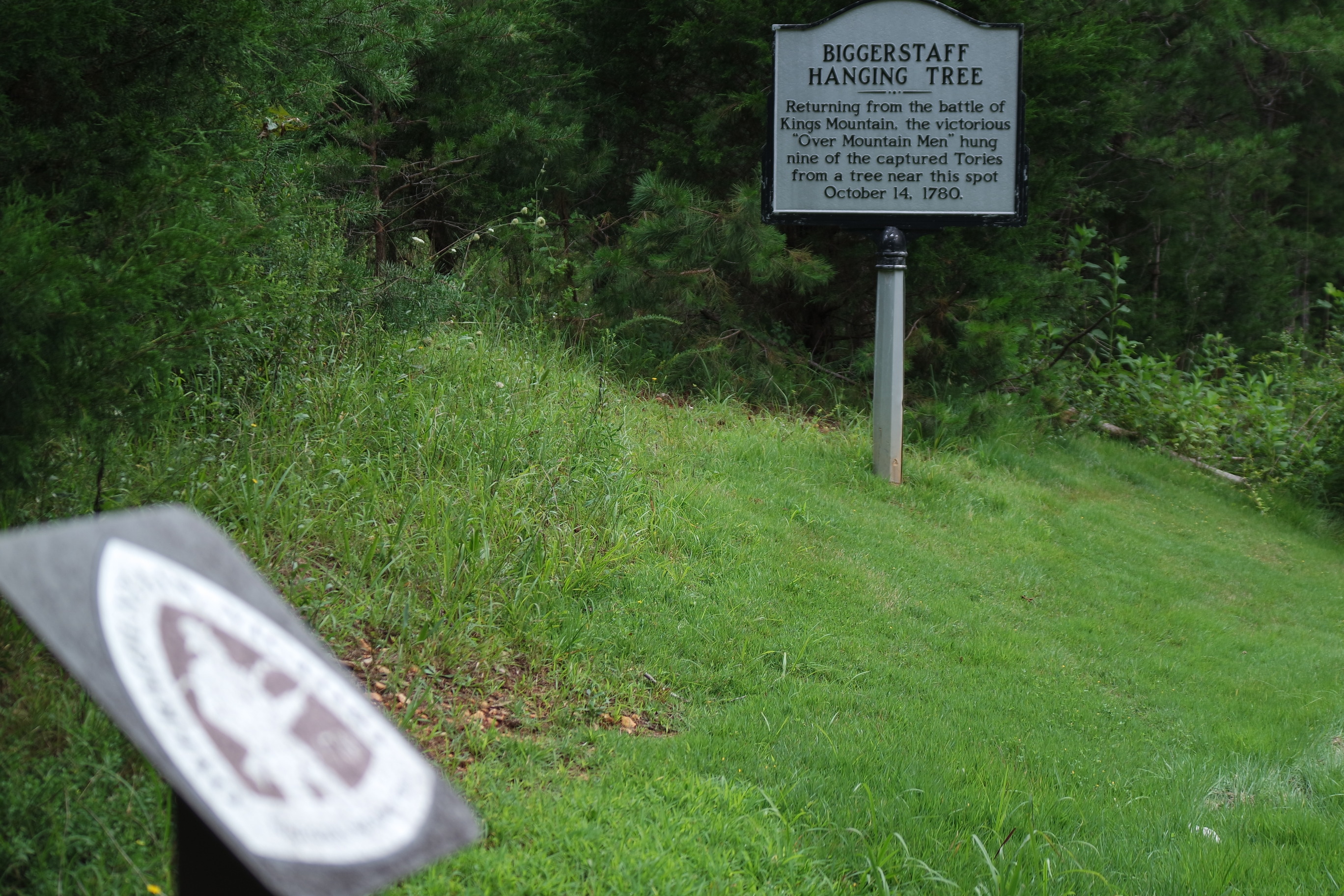 A white metal historical marker with black text reading "Biggerstaff Hanging Tree" stands in the background. In the foreground is a blurry image of the brown metal sign with white triangular logo of the Overmountain Victory National Historic Trail. 