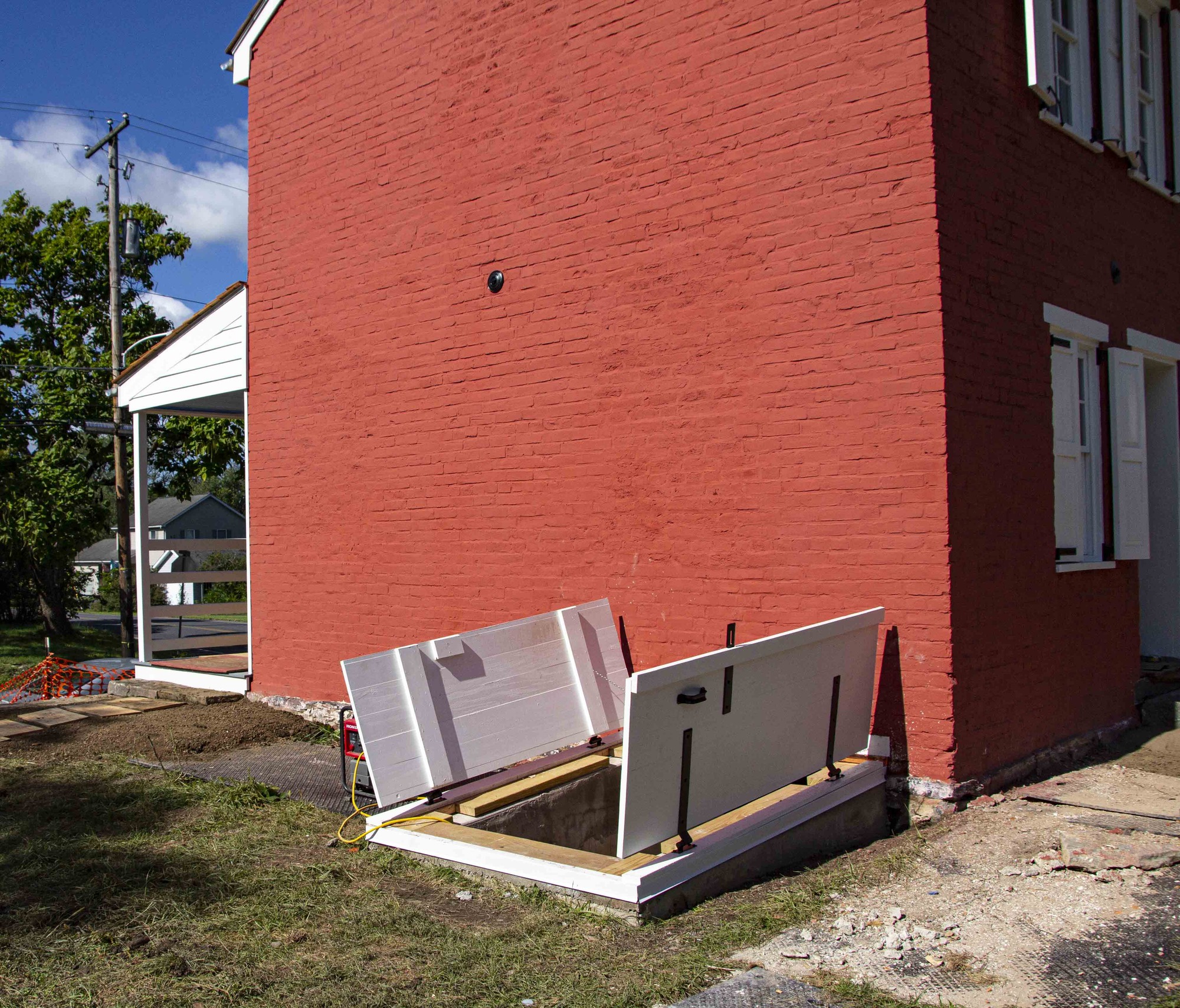 The newly installed storm cellar doors are open leading to the basement of the house. A red generator can be seen beside the entrance storm cellar with a yellow cable leading down into the basement. To the left of the cellar is a newly installed stone walkway that leads to the front porch.