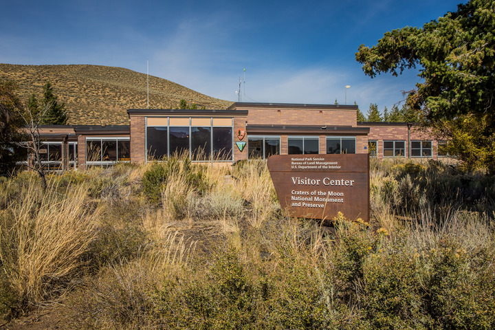 a brick building with large rectangular windows behind a patch of tall grass and shrubs with a sign reading "national park service, bureau of land management, US department of the interior: visitor center, craters of the moon national monument and preserve"