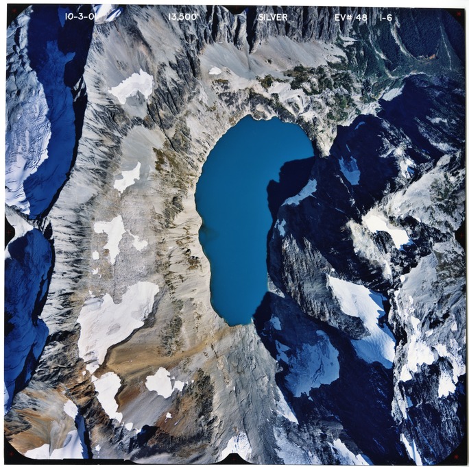 Color aerial photograph of glacier and snowfields on mountain peak with forested valleys below.