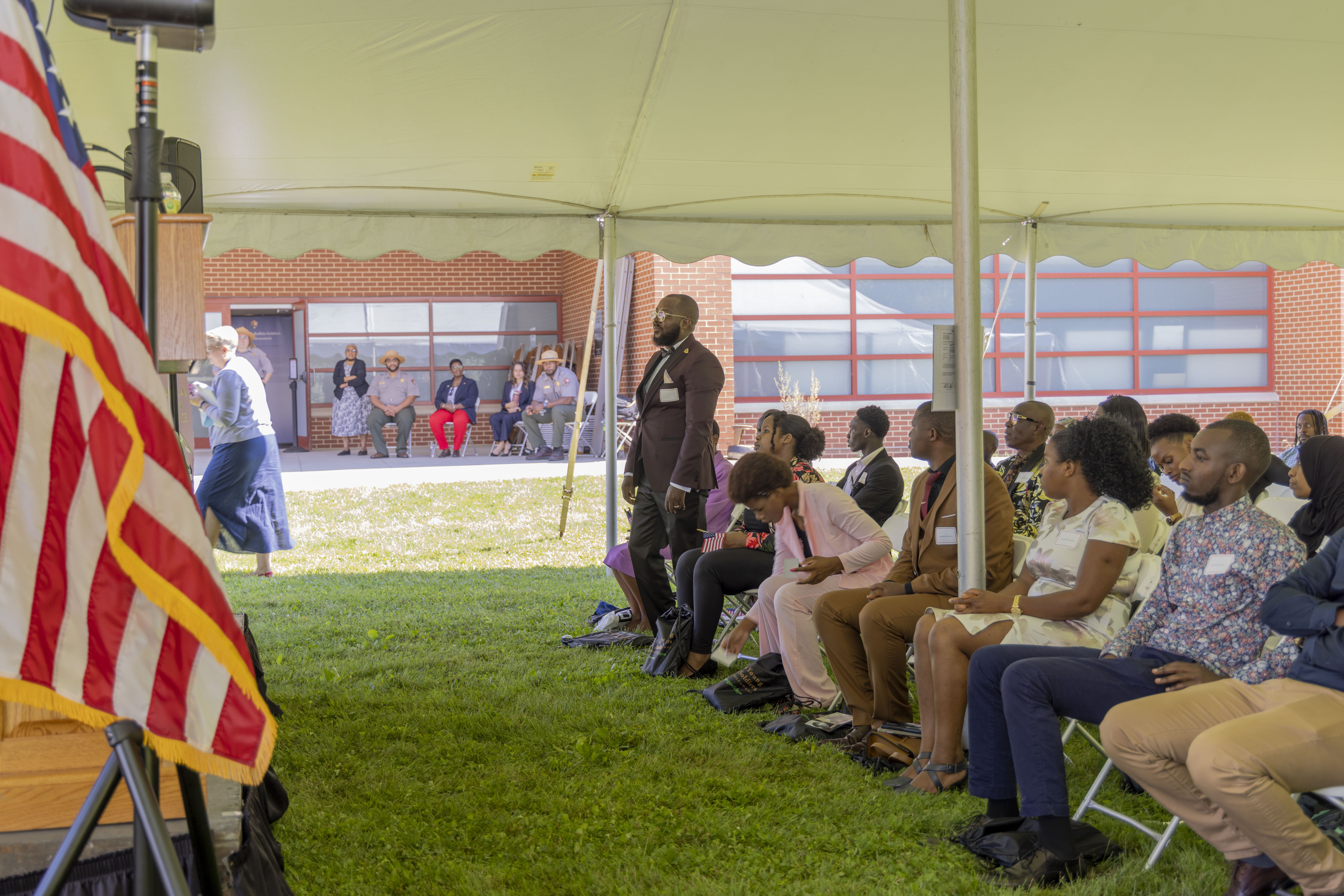 A man wearing a dark-colored suit stands amongst several seated people and speaks to them.