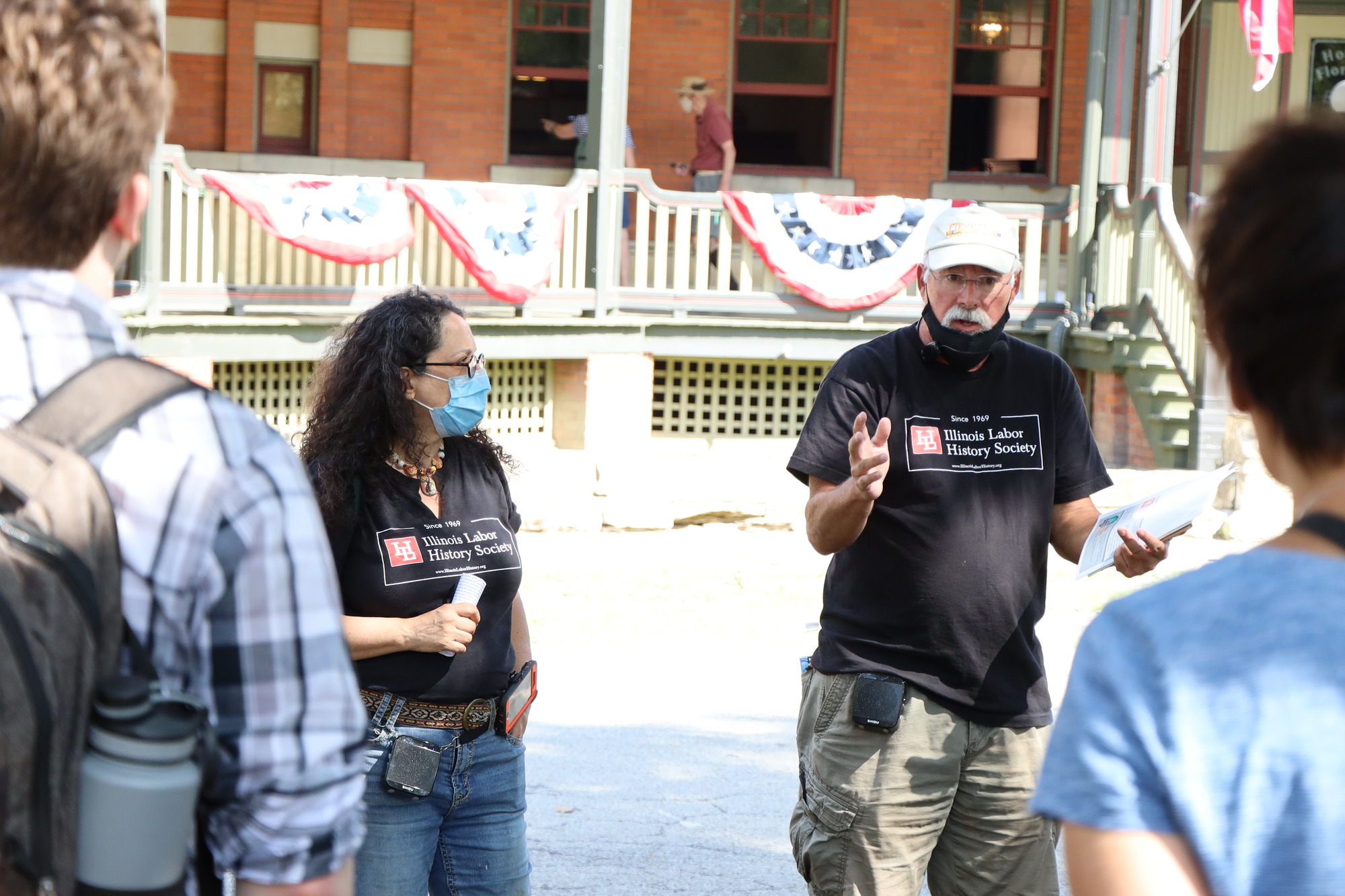 Photo of two tour guides standing in front of the Hotel Florence talking to a group of people. The tour guides wear matching black tshirts that say Illinois Labor History Society with its logo. The tour guide on the left is a Latina woman with long curly hair wearing jeans. The tour guide on the right is an older white man with a white mustache, white cap, and wearing khaki pants. 

The back of two visitors at either side of the photo can be shown looking towards both of the tour guides. 