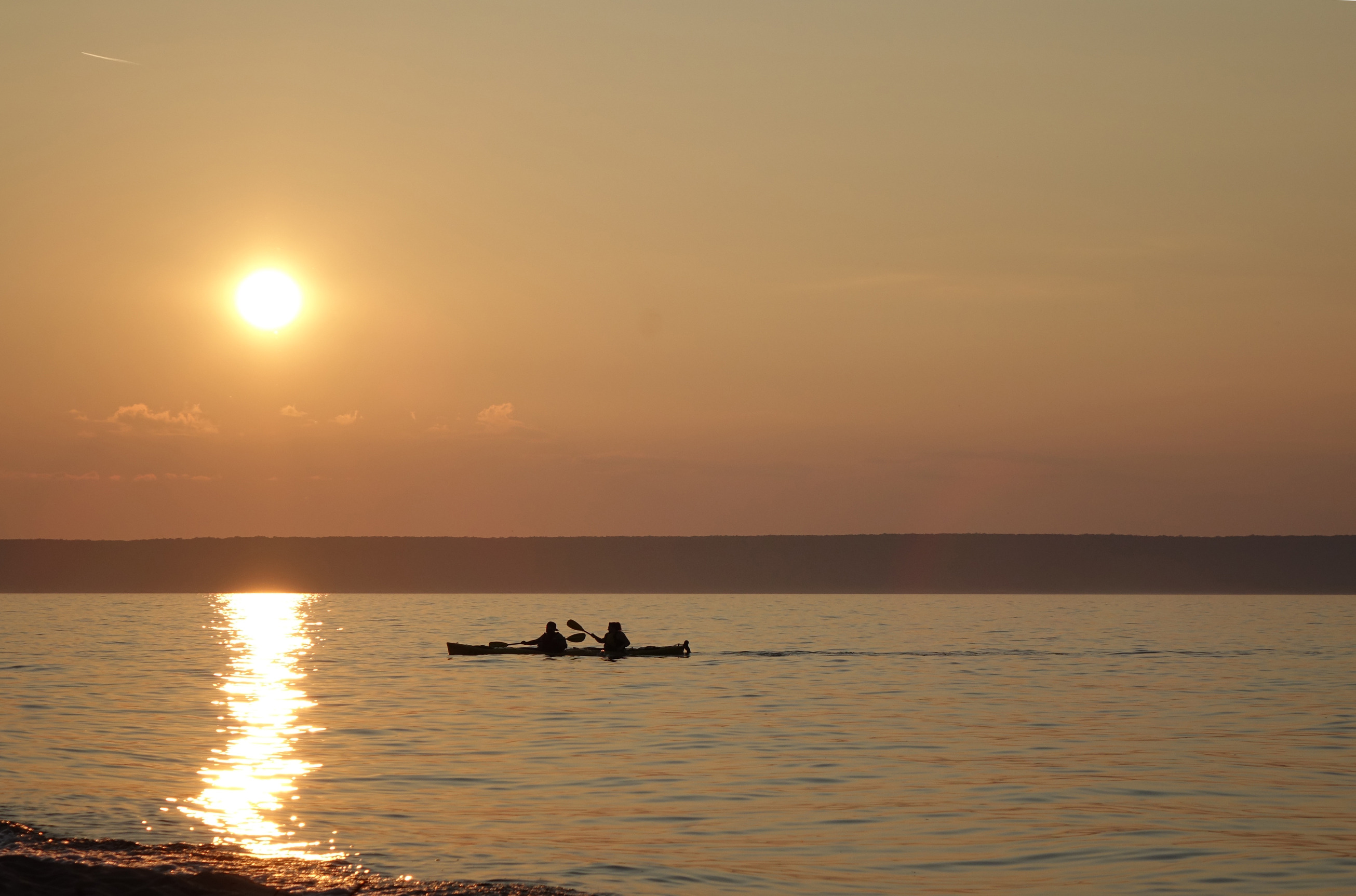 two people in a tandem kayak paddling while the sun is approaching the horizon. The sun is reflecting in a line across the water in front of the kayakers.