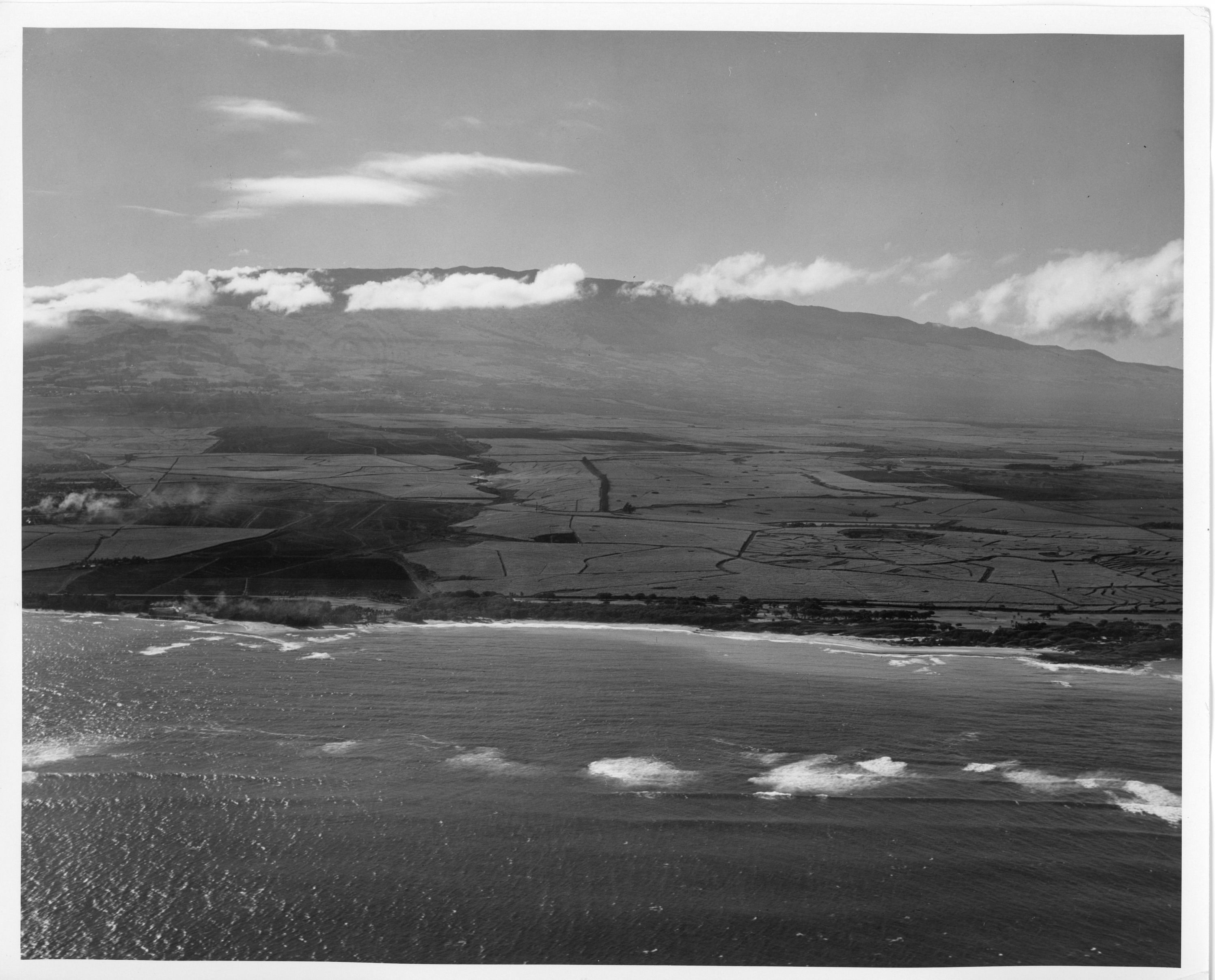 A black and white aerial image of Maui and Haleakalā. The bottom of the image shows the ocean and the waves moving and crashing onto the shoreline. The coast is visible with sand and trees in the distance. Above the trees are the sugar cane fields that cover a large portion of the land. At the very top of the image is Haleakalā volcano. Above the volcano, clouds fill the sky and cover portions of Haleakalā.