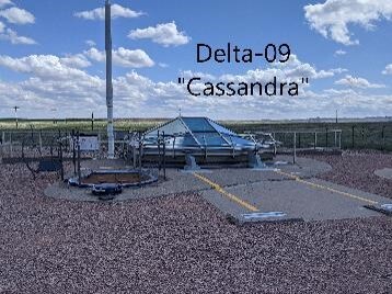 Gravel area with several hatches on a cement pad with a glass dome in the center and a white pole sticking up. Words "Delta-09 Cassandra"