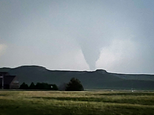 Dark gray funnel cloud descending from clouds over hills and a brown building.