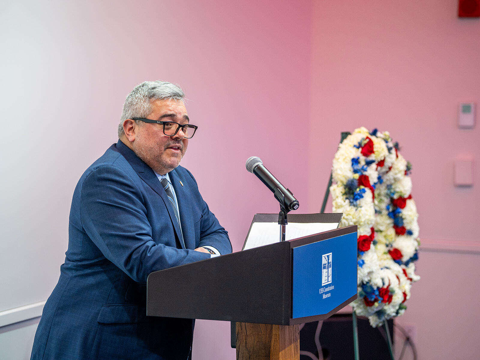 Man in blue suit with glasses stands behind a podium giving remarks.